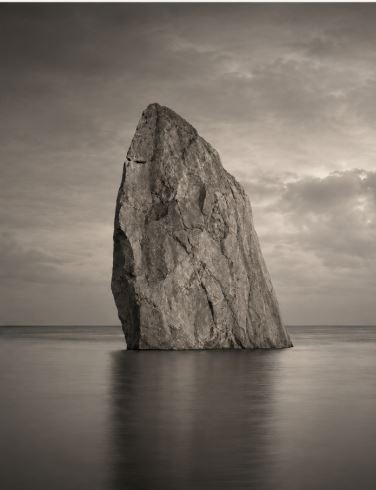 A single monolithic rock rising from still water under soft grey cloud